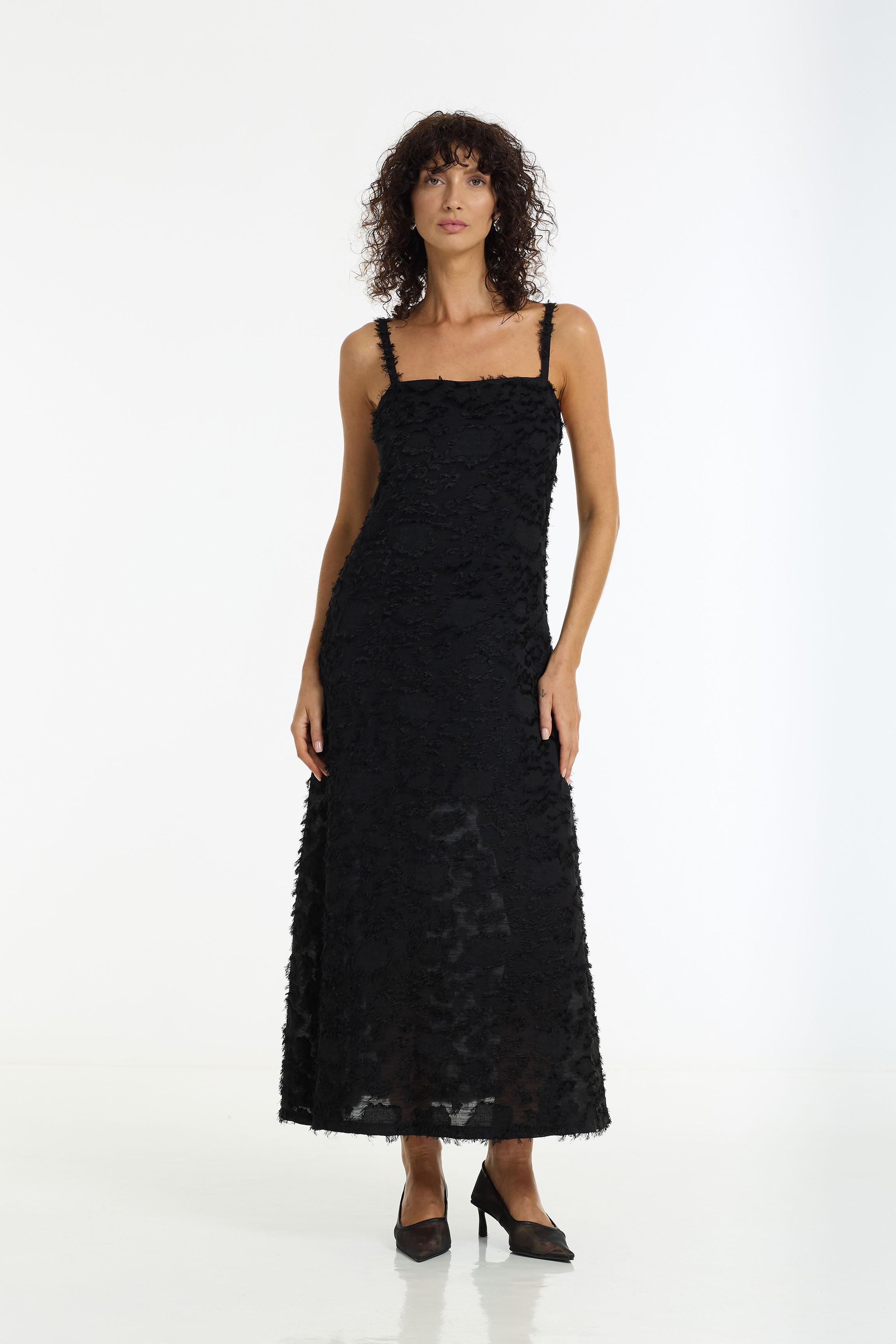 A woman modeling a Wildest Hearts Textured Maxi Dress in black by BLAK, paired with black pointed-toe heels, against a plain white studio background.